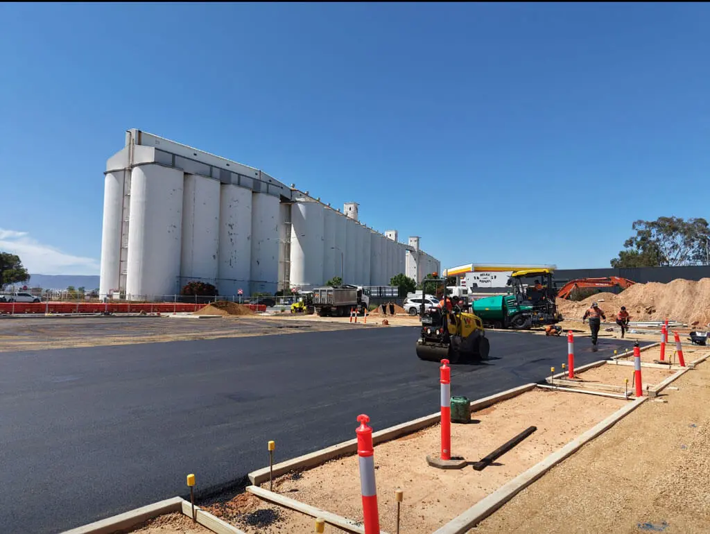 Construction workers and machinery paving a road near large white industrial silos on a sunny day, with orange safety cones and fencing marking the construction area.