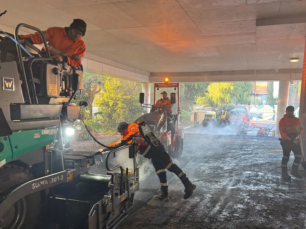 Workers in high-visibility clothing operate heavy machinery and lay asphalt under a bridge, with steam rising from the ground and light streaming in from the background.