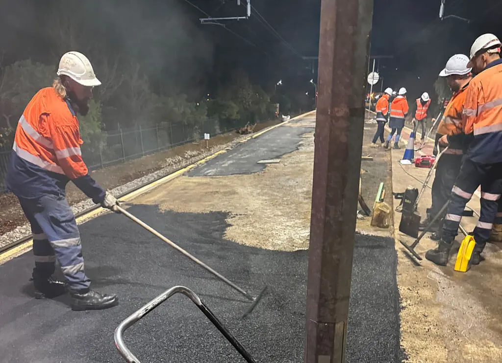 Workers in orange safety vests and helmets lay and smooth fresh asphalt on a railway platform at night, using tools under artificial lighting. Trees and train tracks are visible in the background.