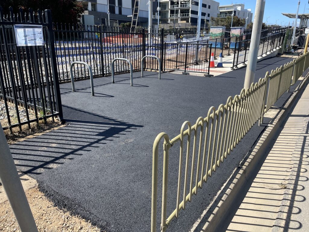 A newly paved black asphalt path leads to bike racks between black metal fences, with yellow railing in the foreground and buildings visible in the background on a sunny day.