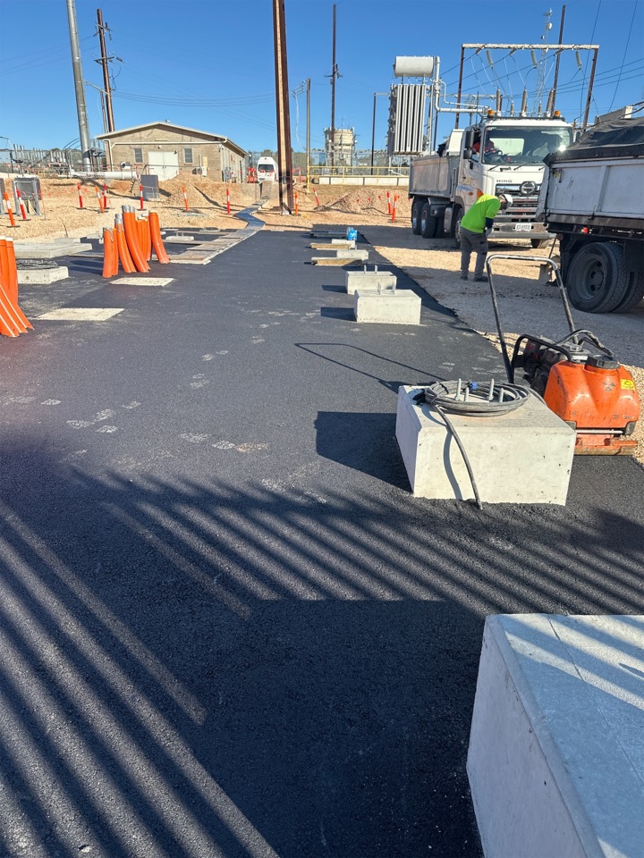Freshly paved asphalt area at an industrial site with concrete blocks, orange safety barriers, construction equipment, and a worker in a green shirt near two large utility trucks under a clear blue sky.