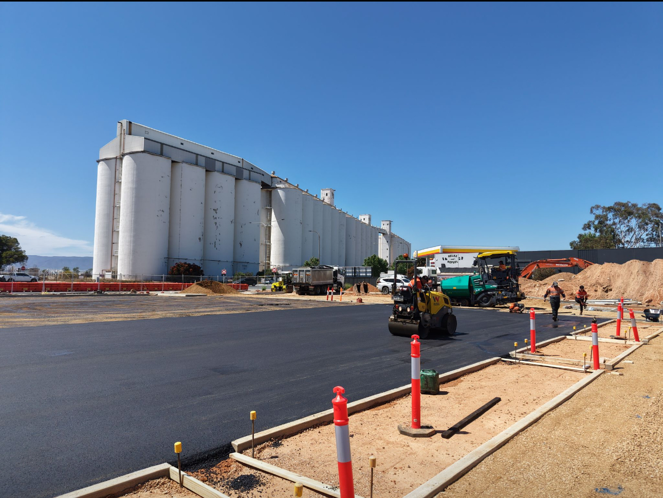 Construction workers and heavy machinery pave a road near large white industrial silos under a clear blue sky. Orange safety barriers and traffic cones line the construction site.