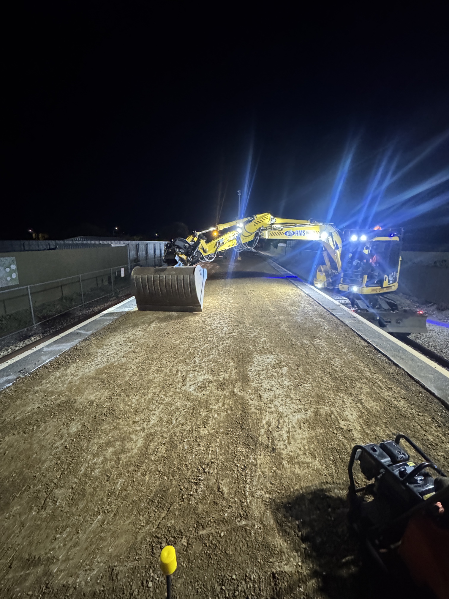 A large yellow excavator works on a dirt construction site at night, illuminated by bright blue and white lights, with construction equipment and materials visible in the background.