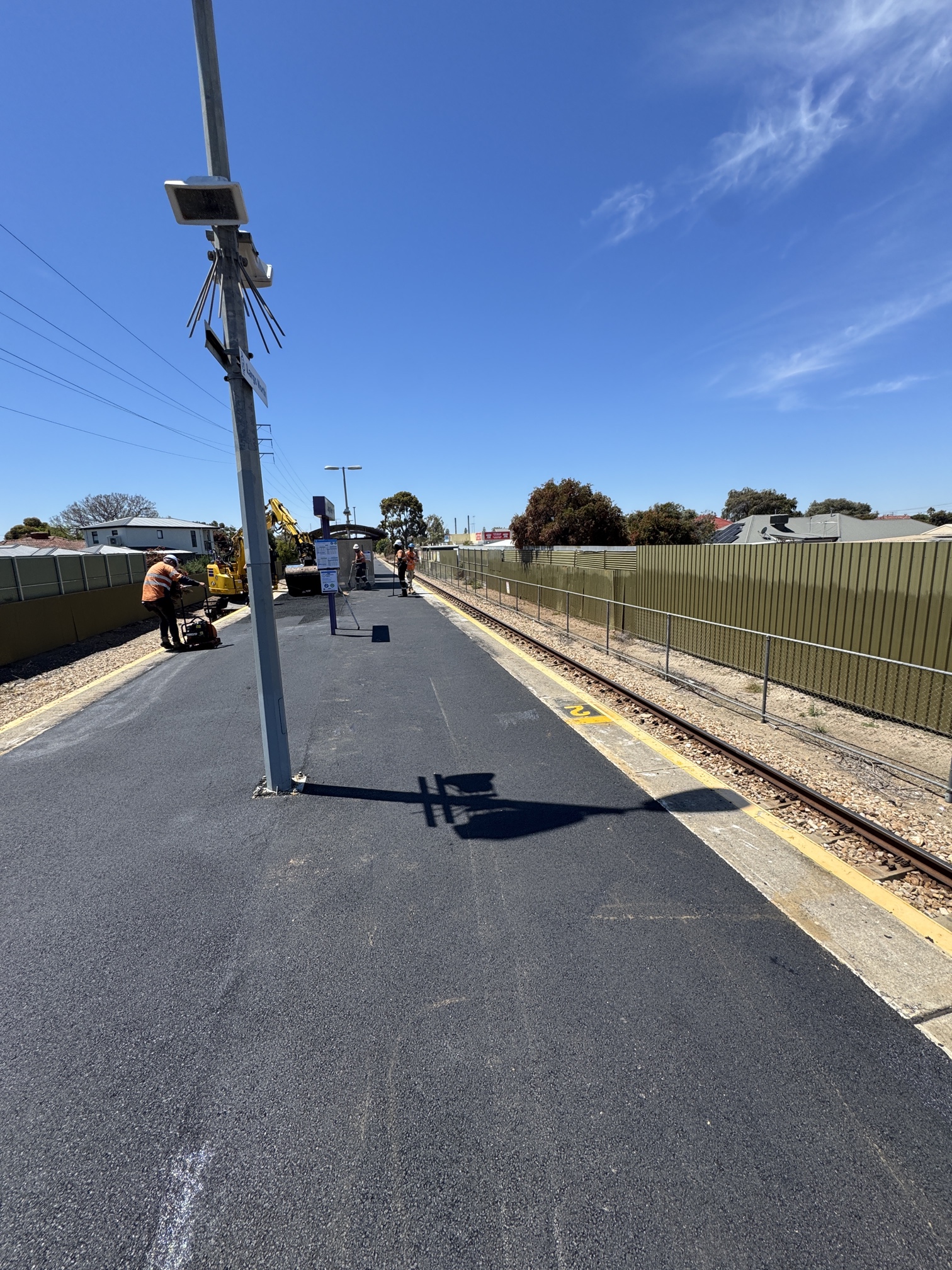 Construction workers are paving a train station platform on a sunny day. Equipment and vehicles are present, with a metal pole in the foreground and train tracks running alongside the platform.