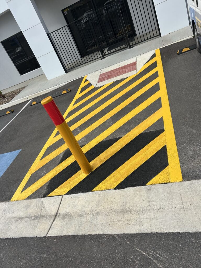 A yellow and black striped no-parking zone is painted on asphalt next to a sidewalk ramp, with a yellow bollard marked red at the top in the center. A building and parking stops are visible in the background.