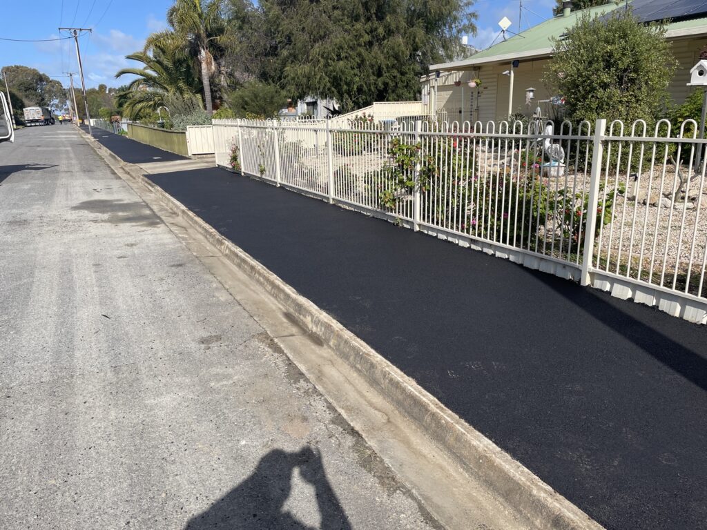 A freshly paved black asphalt footpath runs alongside a white picket fence and a garden in front of a house on a sunny day. A shadow of the person taking the photo is visible on the street.