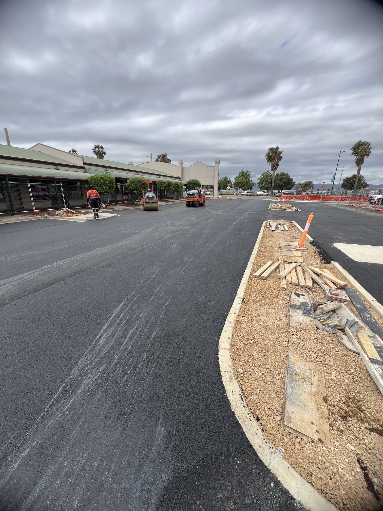 Freshly paved road with construction workers and equipment, bordered by dirt and construction materials on the right side. Buildings and palm trees are visible in the background under a cloudy sky.