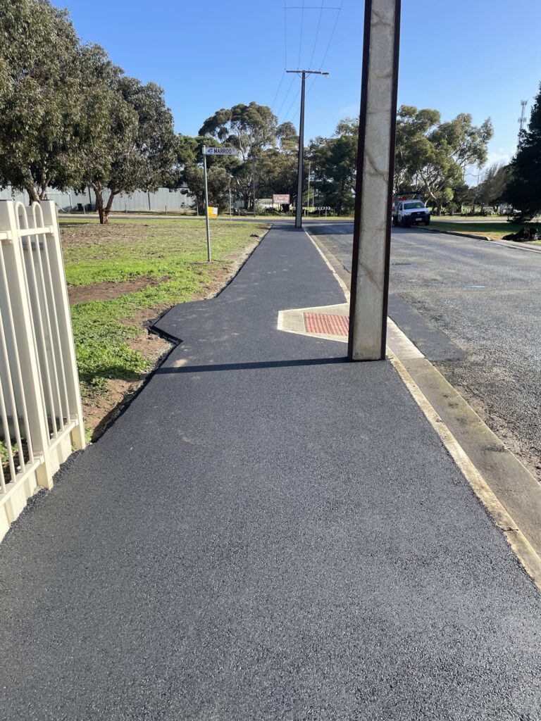 A freshly paved sidewalk with a utility pole obstructing the middle, next to a grassy area and a parked car on the street under a clear blue sky.