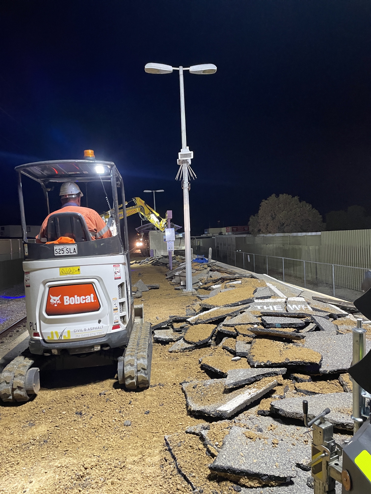 A construction worker operates a small excavator at night, breaking up asphalt on a pathway. Broken pavement pieces are scattered, and another worker and machine are visible ahead under streetlights.