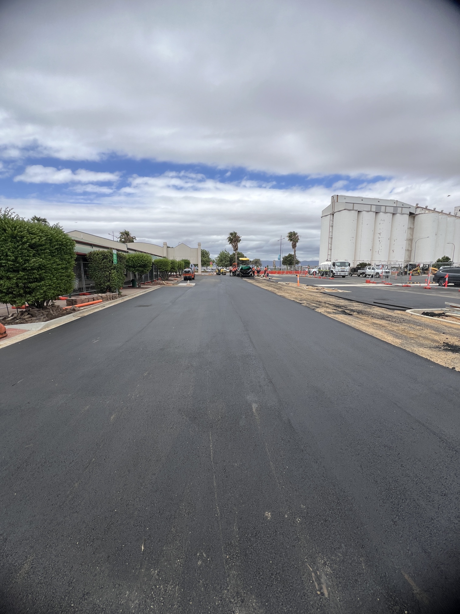 Freshly paved road with new black asphalt, bordered by construction cones and equipment. To the right are tall industrial silos, and there are some trees, bushes, and partly cloudy skies overhead.