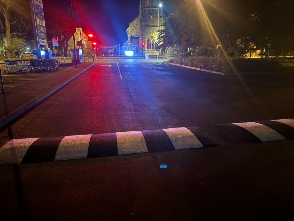 A freshly paved empty parking lot at night, illuminated by streetlights and red traffic lights, with a black-and-white striped speed bump in the foreground and a lit stone church building in the background.