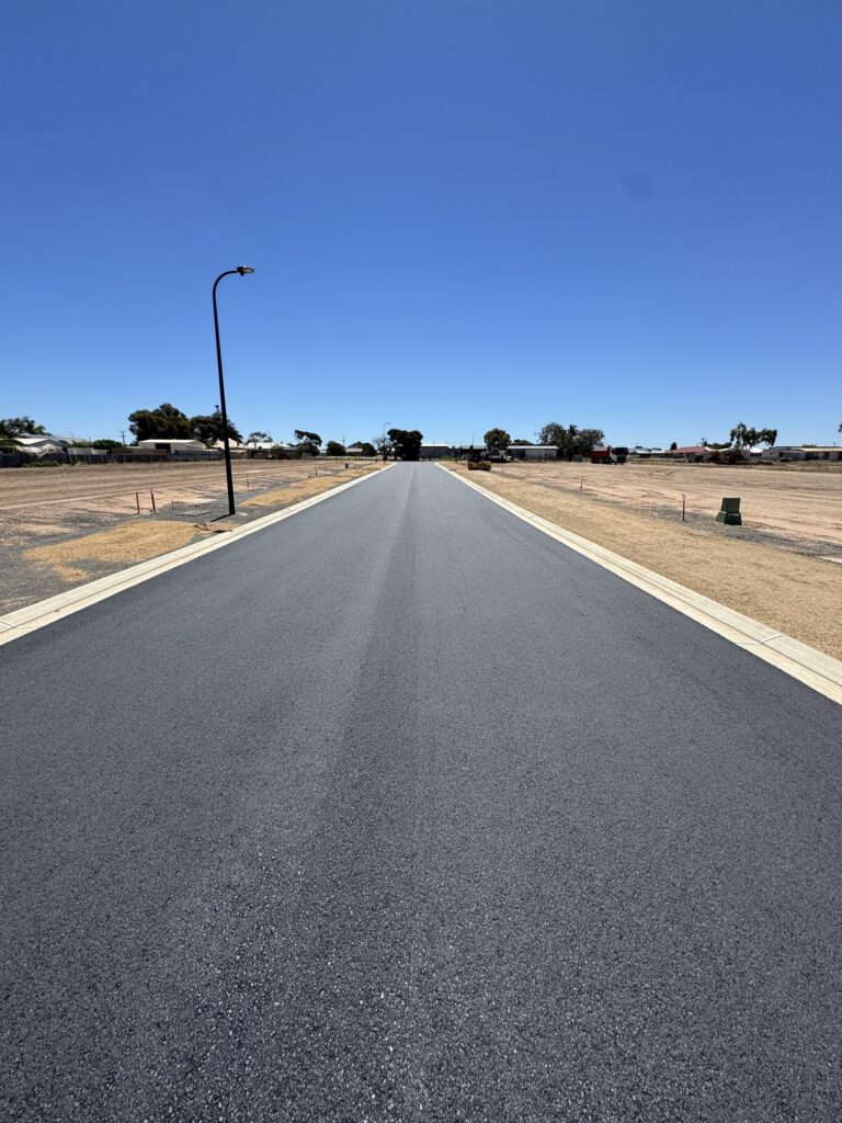 A freshly paved, empty road stretches straight into the distance under a clear blue sky, with bare, dry land and sparse trees on either side. A single streetlight stands on the left.