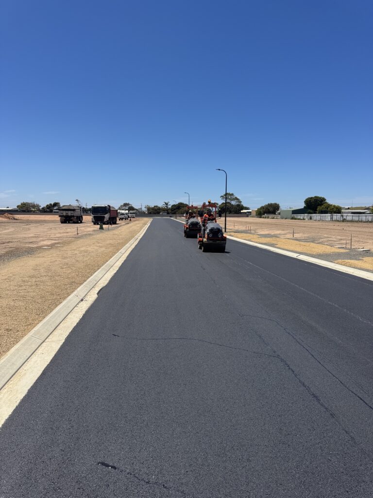 A newly paved road stretches into the distance under a clear blue sky, with construction vehicles and workers visible ahead. Dry, sandy ground lines both sides of the road, and buildings are visible on the right.