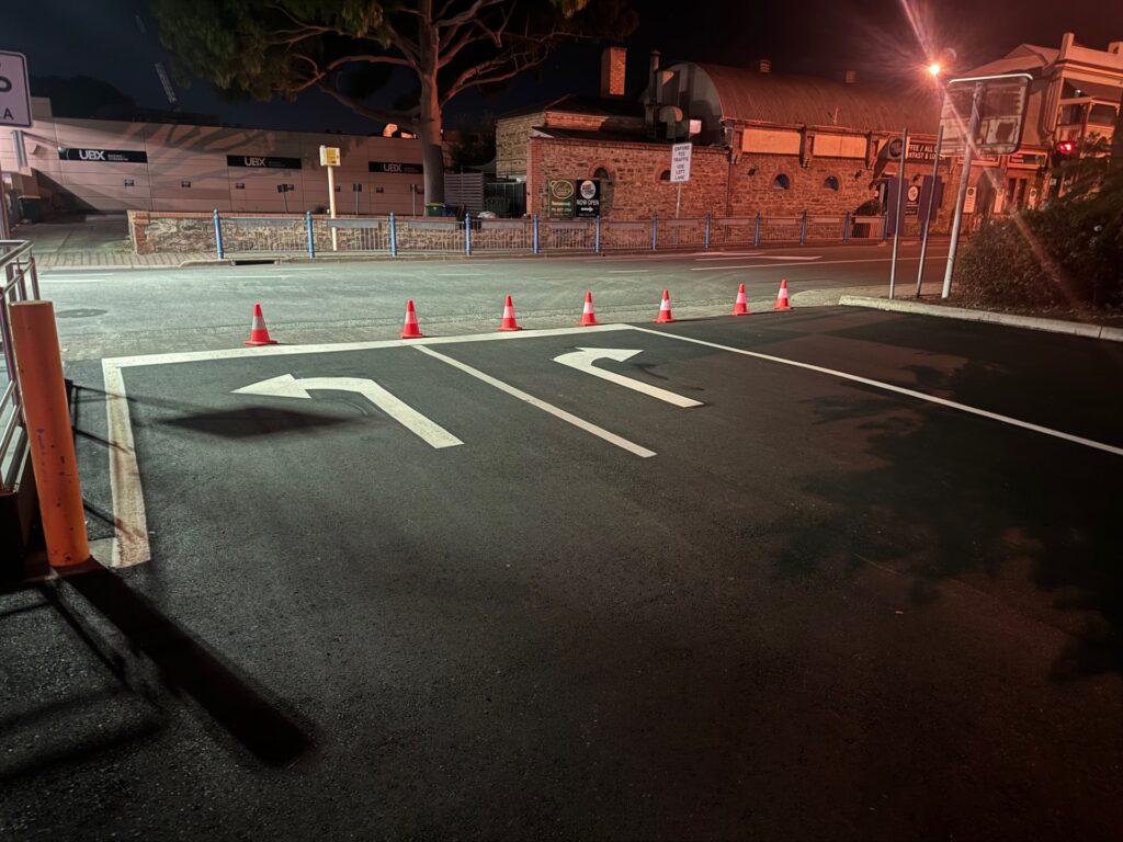 An empty parking lot at night with white arrows on the ground and a row of orange traffic cones blocking entry. Buildings and streetlights are visible in the background.