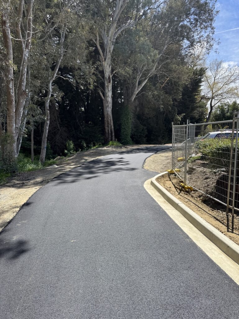 A newly paved asphalt path curves through a wooded area with tall trees on the left and a construction fence with dirt on the right, under a sunny blue sky.