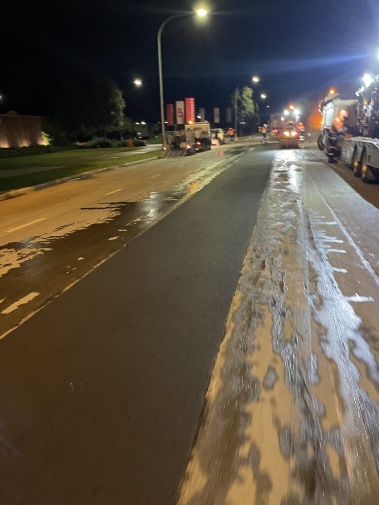 A nighttime road scene shows a freshly resurfaced street with wet, reflective pavement and foam lines. Streetlights illuminate construction vehicles and workers in the distance.
