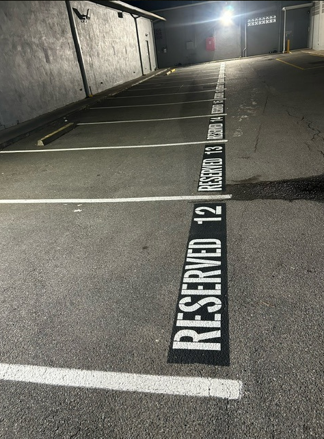 A row of empty parking spaces at night, each marked with a white “RESERVED” label and corresponding numbers painted on the ground. The area is lit by overhead lights.