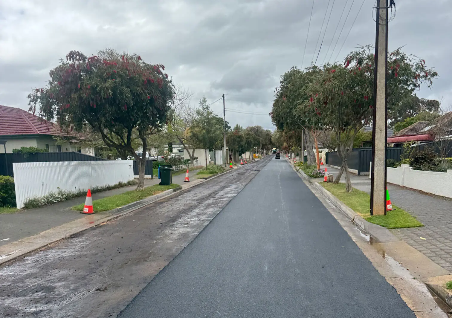 A freshly paved residential street with traffic cones along the sidewalk, trees on both sides, and overcast skies above. Some dirt remains on the edge of the road, indicating recent roadwork.