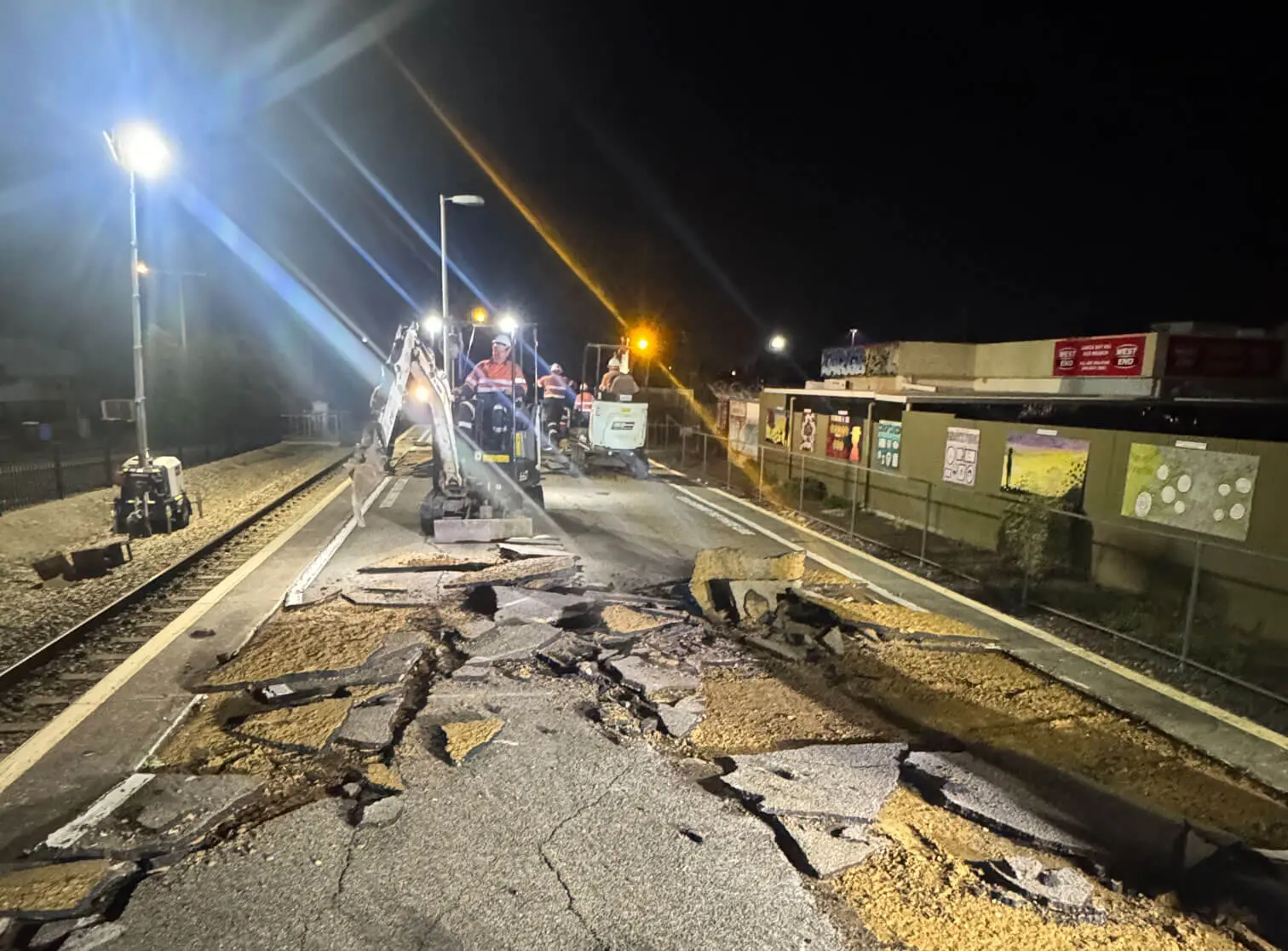 Construction workers operate heavy machinery at night, breaking and removing cracked asphalt on a train platform under bright lights. Debris and large chunks of pavement cover the ground.