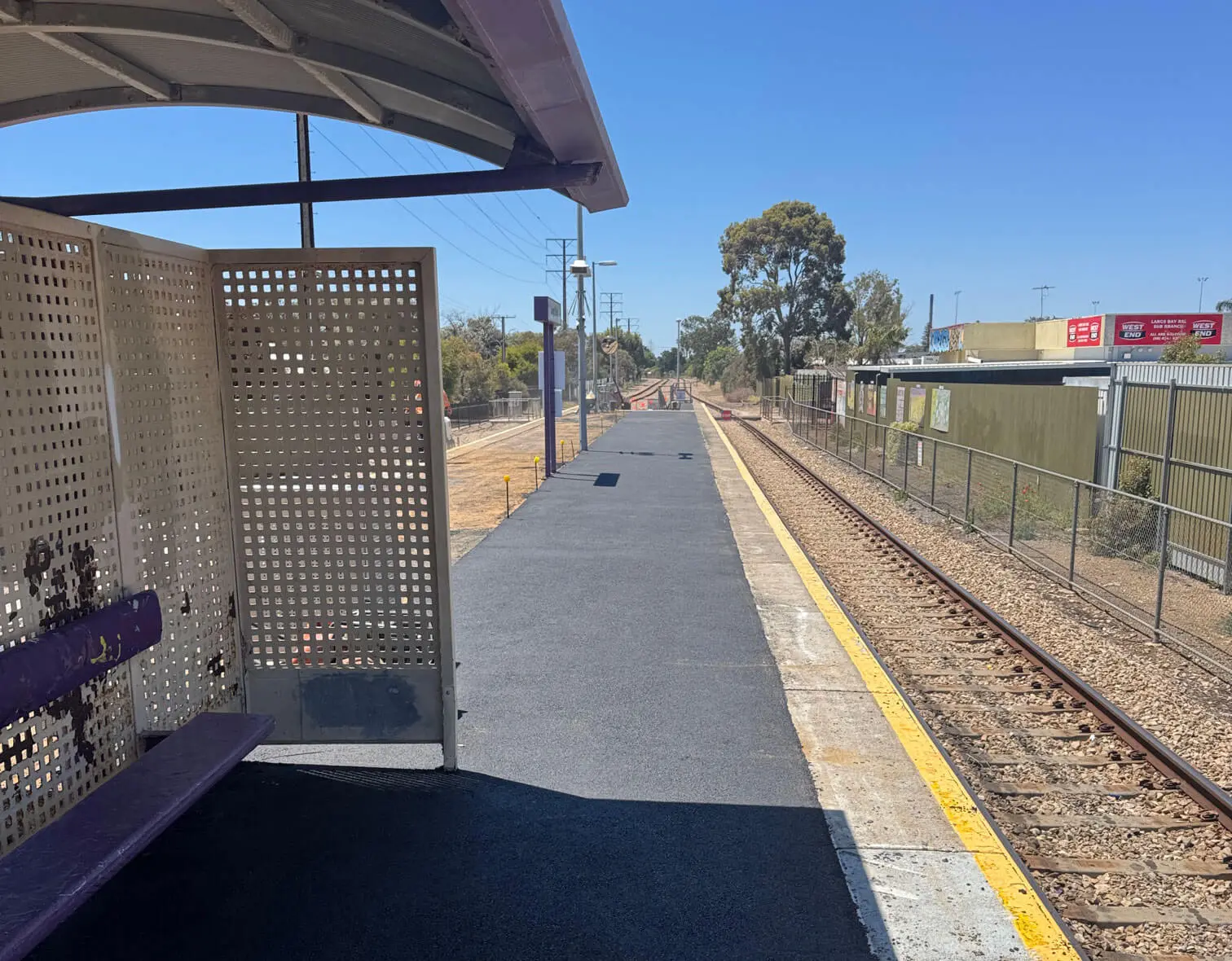 A quiet, empty train station platform with a bench and shelter on the left, train tracks running alongside, and industrial buildings and clear blue sky in the background.