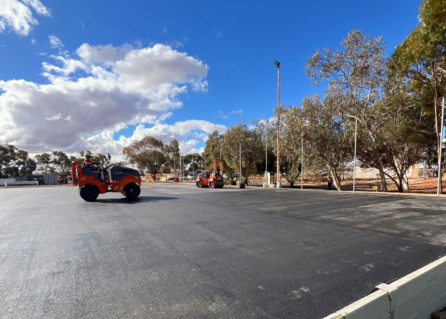 Two construction vehicles work on newly paved asphalt under a partly cloudy blue sky, surrounded by trees and light poles in an open area.