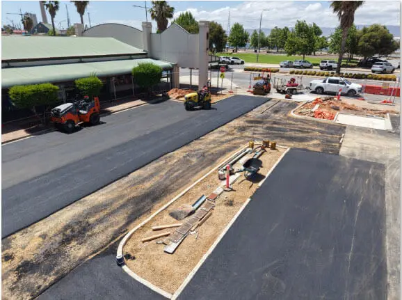 A road construction site with fresh asphalt, construction vehicles, traffic cones, workers, and tools scattered on a median strip. Nearby are buildings, parked cars, and trees under a partly cloudy sky.