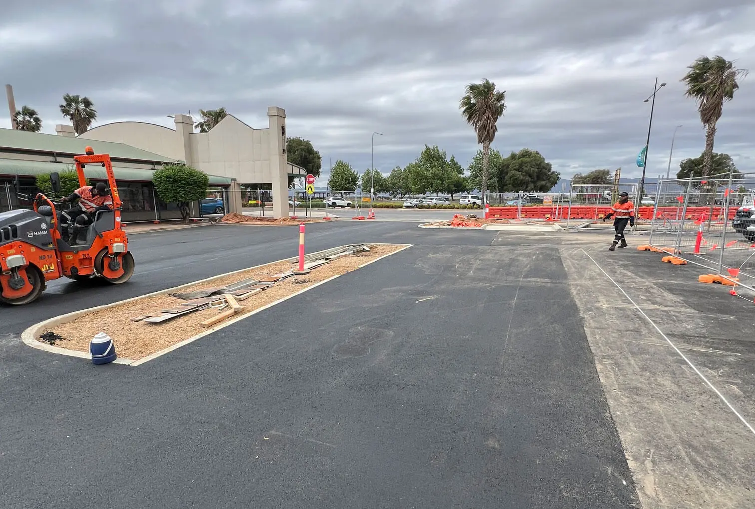 A construction site in a parking lot features fresh asphalt, orange safety barriers, a worker in an orange vest, and a steamroller on the left. There are trees, cloudy skies, and a building in the background.
