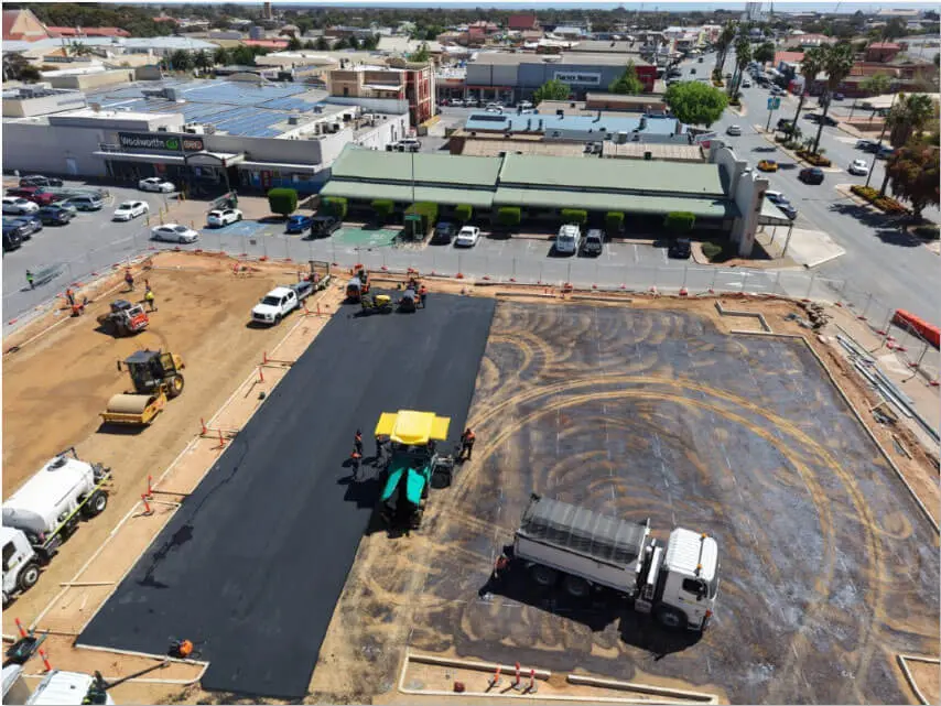 Aerial view of a construction site where workers and vehicles are paving a parking lot with asphalt, adjacent to a supermarket and other shops in a commercial area.