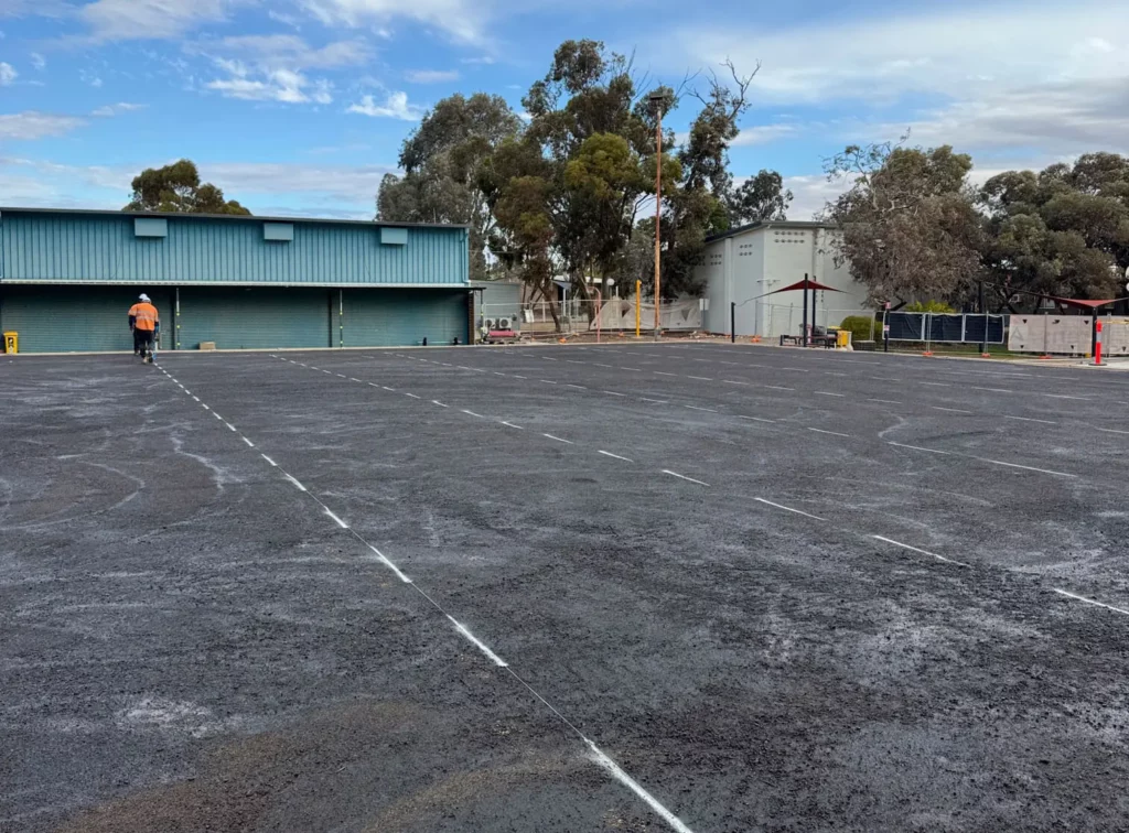 A construction worker wearing an orange vest walks across a large, freshly paved area with white chalk lines, near a blue industrial building and trees under a partly cloudy sky.