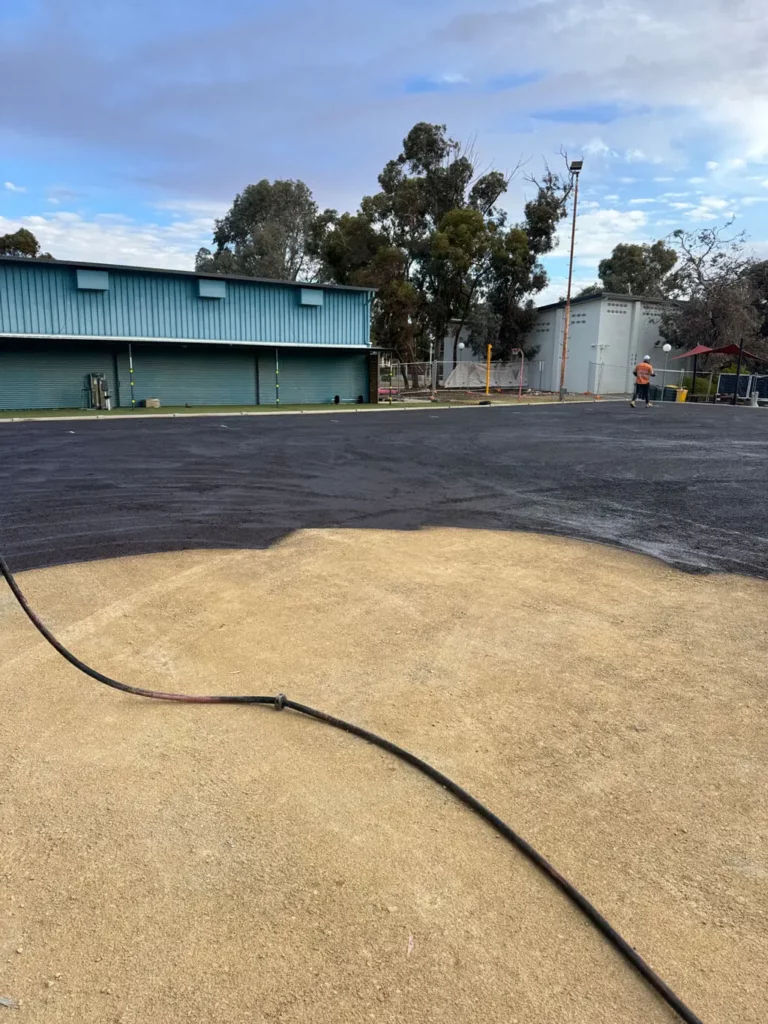 A construction site with freshly laid asphalt next to a dirt area; a hose lies on the ground, and a person in a safety vest stands near industrial buildings and trees under a partly cloudy sky.