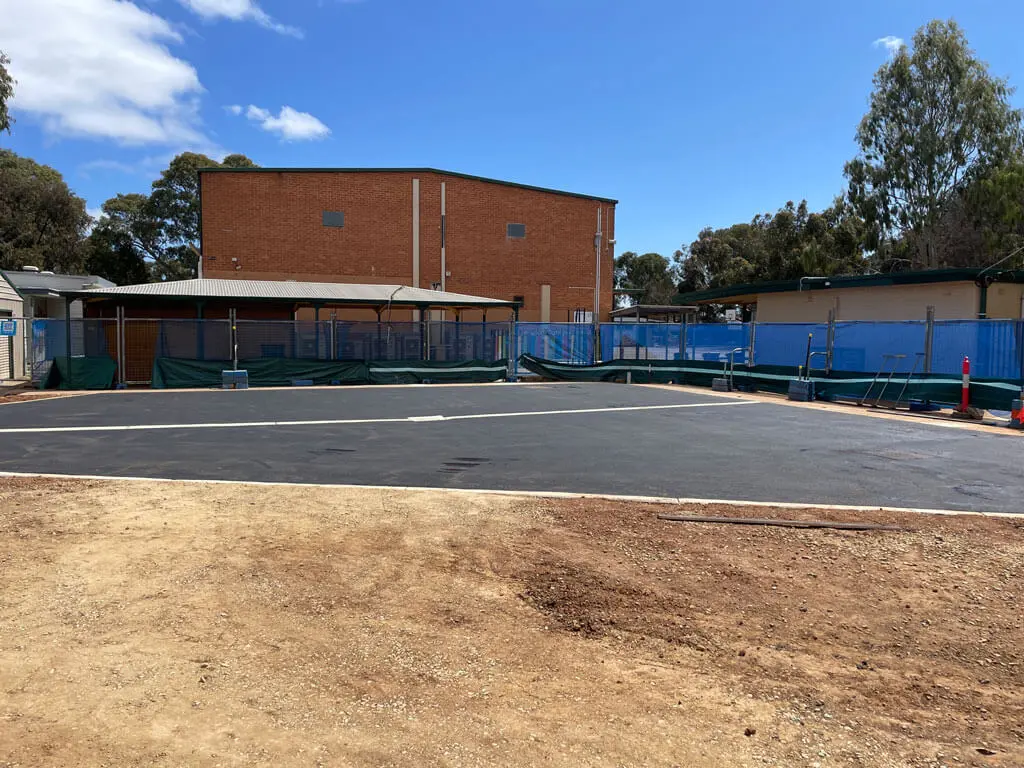 A freshly paved asphalt area surrounded by green and blue construction fencing, with a brick building and trees in the background under a blue sky with some clouds.
