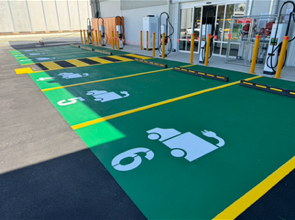 Six green parking spaces with white electric vehicle symbols and numbers, marked for EV charging, are outside a building entrance. Charging stations and yellow safety bollards are visible near the spaces.