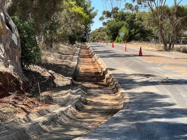 A roadside shows deeply grooved tire tracks cutting through a dirt ditch next to an asphalt road lined with orange traffic cones and trees under a clear sky.