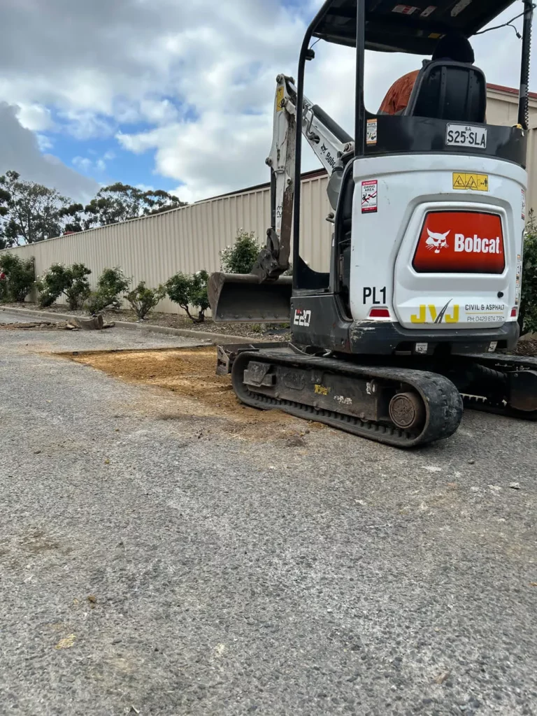 A small Bobcat excavator is parked on a gravel surface near some freshly dug earth, next to a row of trees and a large beige metal fence under a partly cloudy sky.