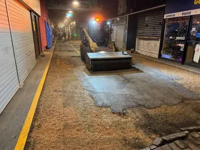 A street cleaning machine sweeps debris on a dimly lit, empty urban street at night. Storefronts with closed shutters line both sides of the street, and overhead streetlights illuminate the scene.
