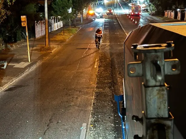 A worker in a reflective vest walks along a dimly lit road at night. Traffic cones, streetlights, and vehicles are visible in the background, while machinery or equipment appears in the foreground on the right.